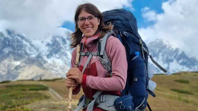 Portrait von Hannah Hütter in roten Pullover und blauen Rucksack, Hintergrund ist ein Weg und Berge